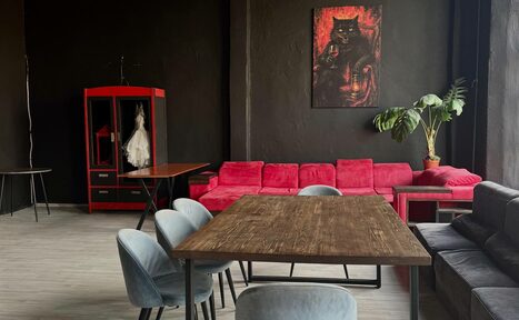 Арендовать C. A. T. Group (C.A.T. Группа). Пpoстоpный двуxэтaжный лoфт. Просторное помещение с черными стенами, деревянным полом. Красный диван, серый стул, деревянный стол. Красная гардеробная с белым платьем. На стене картина с изображением кошки. Растение в горшке, большое окно справа.