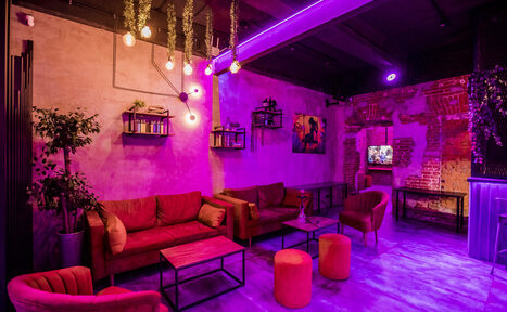 Арендовать Дизайнерский лофт с караоке и активностями возле метро. Лофт-пространство с кирпичными стенами, диванами и креслами красного цвета. На потолке висят лампы накаливания. Есть растения и полки с книгами. Освещение фиолетовое. На стене картина и висят декоративные элементы. В комнате стулья и журнальные столики.