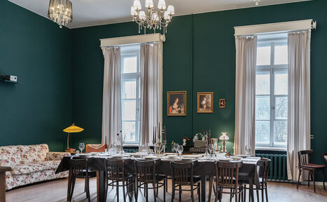 Арендовать Пространство Имена. Винтажный лофт . Просторная комната с тёмно-зелёными стенами и белым потолком. Большой стол, сервированный для ужина, окружён деревянными стульями. Справа — две высокие окна с белыми шторами. На стене — картины. Люстра и настольные лампы обеспечивают освещение.