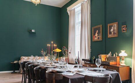 Арендовать Пространство Имена. Винтажный лофт . Столовая с зеленым интерьером, длинным столом и черными стульями. На столе белая посуда и бокалы. На заднем плане диван, комод с лампами и картинами. Высокие окна с белыми занавесками. Светильник на потолке.