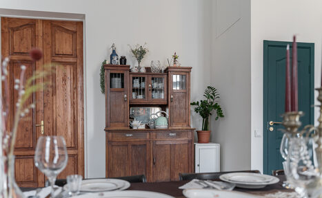 Арендовать Пространство Имена. Винтажный лофт . Столовая комната с деревянным буфетом, обставленным посудой и декоративными элементами. На переднем плане накрытый обеденный стол с тарелками и бокалами. Светлые стены, деревянные двери, зелёное растение в углу.
