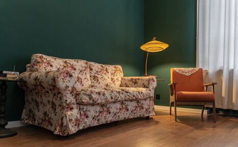Арендовать Пространство Имена. Винтажный лофт . Гостиная с зелеными стенами, на полу деревянное покрытие. Диван в цветочной обивке, рядом оранжевое кресло. Торшер с абажуром, белые шторы на окне. Небольшой стол с книгой и лампой.