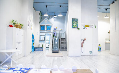 Арендовать Лофт Облака. Зал Баунти на Полярной. Детская игровая комната с деревянными полами и белыми стенами, оформленная в светлых цветах. Возвышение с лесенкой и горкой, декоративный домик, наклейка с жирафом на двери. Стол с белыми стульями и цветной скатертью, зелёные растения на полках.