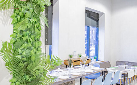 Арендовать Лофт Облака. Зал Баунти на Полярной. Просторное светлое помещение с длинным столом, сервированным тарелками и бокалами, окруженным голубыми стульями. На стене зеленая растительность. Два больших окна с видом на улицу. Нейтральные стены, натуральное и искусственное освещение.