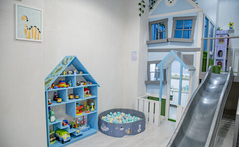 Арендовать Лофт Облака. Зал Баунти на Полярной. Детская комната с горкой, домиком с окнами, игрушечной мебелью, синими стеллажами с игрушками и пластиковым бассейном с разноцветными шарами. Белый пол, светлые стены, теплое освещение. Картина с изображением жирафа на стене.