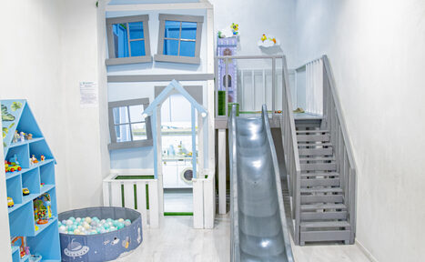 Арендовать Лофт Облака. Зал Баунти на Полярной. Детская игровая зона с деревянными конструкциями в форме домика. Серебристая горка и серые лестницы. Синий стеллаж слева с игрушками. Круглый бассейн с шариками. Светлые стены и пол, бело-голубая цветовая гамма.