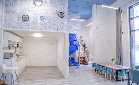 Арендовать Лофт Облака. Зал Баунти на Сельскохозяйственной. Светлое просторное помещение с высокими потолками, кухней слева, серыми шкафами и техникой. Справа обеденный стол с голубыми стульями, рядом лестница и синий винтовой спуск. Светлые стены, большие окна, серый ламинат.