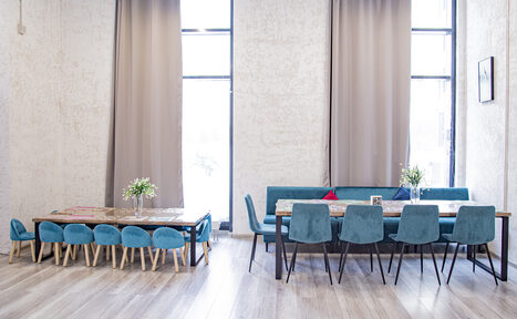 Арендовать Лофт Облака. Зал Баунти на Сельскохозяйственной. Просторное помещение с высокими потолками, двумя большими окнами с бежевыми шторами. Два деревянных стола с сине-зелеными стульями. На столах букеты цветов. Пол светлый деревянный. Стены светло-серые. Справа в верхней части видна рама с картиной.