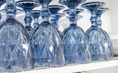 Арендовать Лофт Облака. Зал Баунти на Сельскохозяйственной. Несколько синих стеклянных бокалов перевёрнуты вверх дном на белой полке. Бокалы имеют геометрический узор. Поверхность полки выглядит текстурированной. Видна часть зелёного листа внизу.