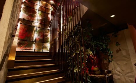 Арендовать Микс Afterparty(После вечеринки). Лестница с металлическими перилами, ведущая вверх. Витражное окно с абстрактным рисунком и подсветкой. Справа декоративное растение в вазе на столике. Стены светлые с текстурной отделкой. Нейтральное освещение.