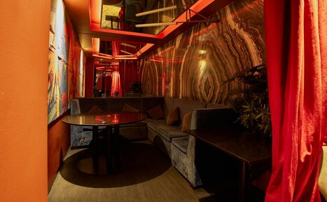 Арендовать Микс Afterparty(После вечеринки). Уютное помещение с полукруглым серым диваном и круглым столом. Стены с мраморной текстурой, оформленные в коричневых и золотистых тонах. Красное неоновое освещение, яркие красные шторы, ковролин теплого оттенка, картины на стенах.