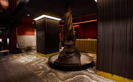 Арендовать Микс Afterparty(После вечеринки). Пространство с круглой скульптурой лошади, отделанной бронзой. Пол мраморный с узором, темные панели на стенах и колоннах, потолочное освещение. Слева кресло и металлический стол. Интерьер в темной и красной гамме.