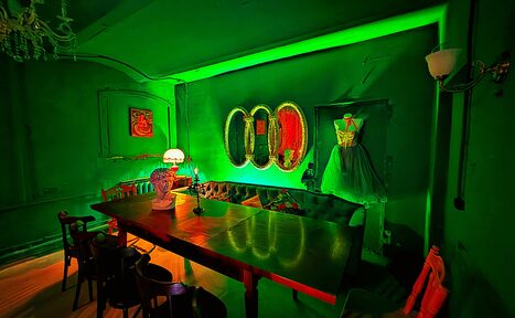 Арендовать Лофт «Рич» (RICH). Комната в зеленых тонах с длинным деревянным столом и стульями. На стене три овальных зеркала, манекен с платьем, картина. На столе настольная лампа и ваза. Потолочный светильник и бра на стене. Пол светлого дерева, стены и потолок зеленые.