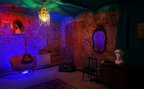 Арендовать Лофт «Рич» (RICH). Подвальное помещение с кирпичными стенами, креслом, гитарой и бюстом на комоде. Винтажное зеркало, деревянный стул и ковёр. Потолочный светильник. Освещение разноцветное, приглушенное. На стенах картины.