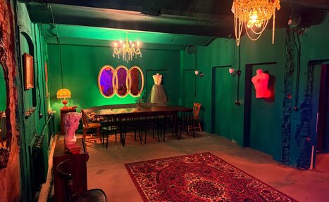 Арендовать Лофт «Рич» (RICH). Комната с зелёными стенами и потолком, длинный деревянный стол с разными стульями, на манекене платье, несколько бюстов, декоративные зеркала, люстра и настольная лампа. Пол покрыт красным ковром с узором. Освещение теплое и разноцветное.