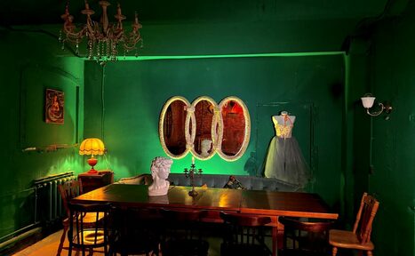 Арендовать Лофт «Рич» (RICH). Затемненное помещение с зелеными стенами. Большой деревянный стол окружен стульями. На столе бюст. На стене овальные зеркала и картина. В углу манекен с платьем. Люстра и настольная лампа обеспечивают освещение.