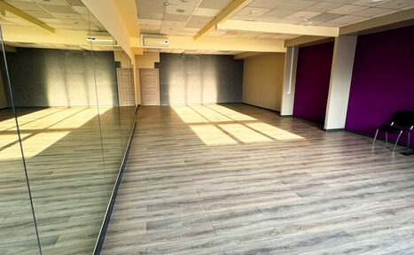 Арендовать Зал №10 «Сеть» 120 кв.м.. Просторное помещение с деревянным полом, зеркальной стеной слева, светлыми потолочными панелями с освещением. Одна стена окрашена в фиолетовый цвет, вдоль неё стоят два стула. Свет от больших окон падает на пол.