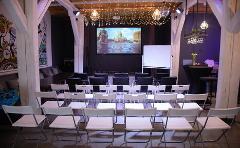 Арендовать Сатурн Лофт. Зал с деревянными балками и видом столов расположенными в форме аудитории. Экран для проекции, флипчарт, подвесные светильники, светодиодные гирлянды. Черные и белые стулья. Слева арт-шаблон на стене. Темно-серые и светло-серые тона интерьера.