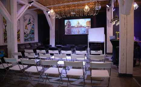 Арендовать Сатурн Лофт. Пространство с креслами, расположенными рядами перед экраном с изображением. Справа флипчарт, стойка с барными стульями. Деревянные балки и потолок. Настенные картины, гирлянды и подвесные лампы. Светлая и темная отделка, серые и белые тона.