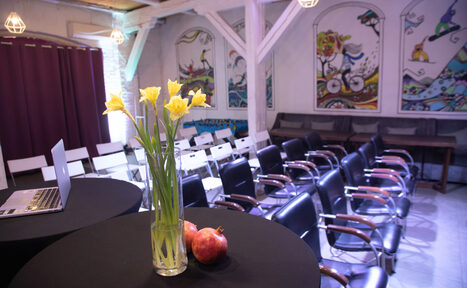 Арендовать Сатурн Лофт. Конференц-зал с креслами в нескольких рядах, столы с черными скатертями, ноутбук, ваза с желтыми цветами, гранаты, на стенах яркие настенные рисунки, фиолетовые занавески, мягкое освещение, балки на потолке.