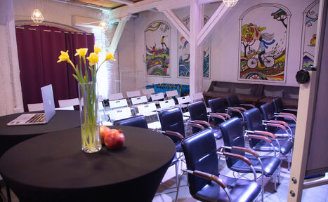 Арендовать Сатурн Лофт. Конференц-зал с рядами черных и белых стульев с деревянными подлокотниками. Стены украшены яркими настенными рисунками, потолок с деревянными панелями. В помещении высокий стол с черной скатертью, вазой с цветами и ноутбуком. Мягкое искусственное освещение.