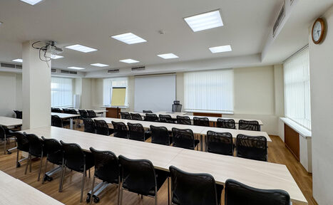 Арендовать Новое образование. Учебная аудитория. Аудиторное пространство с длинными светлыми столами и черными стульями на колесиках в три ряда. Белый экран, проектор и флипчарт. Потолочные светильники, окно с жалюзи, деревянный пол. Светло-бежевые стены, часы на стене.