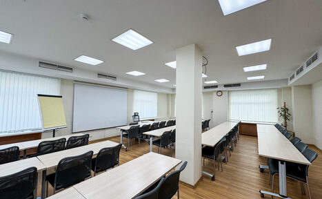 Арендовать Новое образование. Учебная аудитория. Учебное пространство с рядами столов и стульев, проекционный экран, флипчарт и растение. Деревянные полы, светлые стены, потолочные светильники. Вентиляционные решетки и часы на стене. Панорамные окна с вертикальными жалюзи.