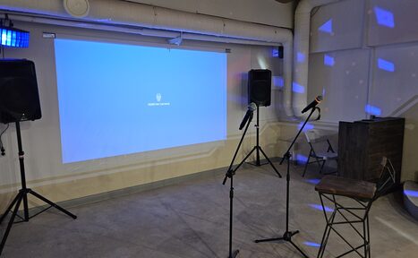 Арендовать Караоке-лофт с бильярдом. Помещение с бетонным полом и светлыми стенами. Проектор проецирует синий экран на стене. Два стула и стойки с микрофонами. Две колонки на стойках. Видны вентиляционные трубы на потолке. Светодиодные светильники создают узоры на стенах.