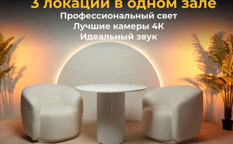 Арендовать Лофт Восторг. Современное помещение с двумя белыми креслами и круглым столом на фоне круглого светящегося элемента. Слева и справа от мебели пальмы в горшках. Текст на изображении: "3 локации в одном зале. Профессиональный свет, Лучшие камеры 4К, Идеальный звук."