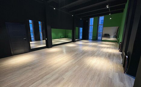 Арендовать Танцевальный зал 80 м.кв.. Просторное помещение с деревянным полом, черными стенами и зелеными акцентами. Зеркальная стена слева, высокие окна справа и на задней стене. Черная дверь слева. На заднем плане стулья, освещение с потолка.