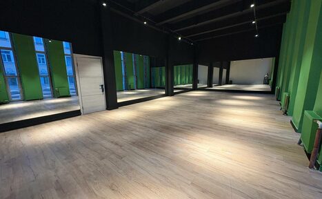 Арендовать Танцевальный зал 80 м.кв.. Просторное помещение с деревянным полом, стены в зелёных и тёмных оттенках, большие окна с отражением домов, светодиодное освещение на потолке, белая дверь сбоку.