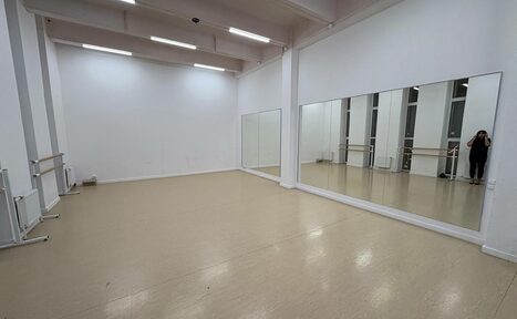 Арендовать Танцевальный зал 60 м.кв.. Просторный зал с бежевым полом, белыми стенами и высокими потолками. Большое зеркало на одной из стен. Установлены светильники на потолке. Справа видны окна и несколько хореографических станков.