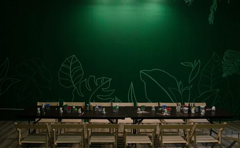 Арендовать Хендмейдщики. Лофт в лесной тематике. Пространство с длинным черным столом и деревянными стульями, расположенными в ряд. На столе множество канцелярских принадлежностей. Задняя стена зеленая с тонкими рисунками листьев. Потолочное освещение с тремя светильниками. Пол светлый.