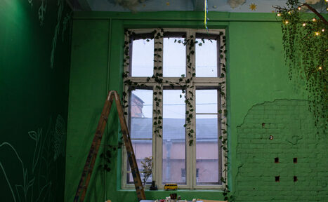 Арендовать Хендмейдщики. Лофт в лесной тематике. Зеленая комната с высоким окном, украшенным искусственными растениями. Стены окрашены в зеленый, частично с обнаженной кирпичной кладкой. В центре деревянный стол с предметами. Деревянная лестница, потолок светло-голубой с облаками и звездами.