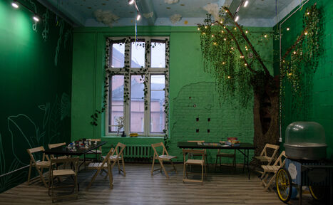 Арендовать Хендмейдщики. Лофт в лесной тематике. Просторное помещение с тёмно-зелёными стенами и деревянным полом. Деревянные складные стулья расставлены вокруг чёрных столов. На стене декоративное дерево с гирляндой. Большое окно с растениями, радиатором. Слева от окна аппарат с прозрачной крышкой. Подвесные светильники.