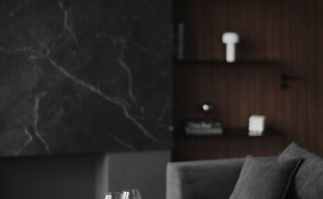 Арендовать Mimika(Мимика). Просторная фотостудия. Современная гостиная с темной мраморной стеной, коричневой деревянной панелью и полками. На фоне темный диван, на переднем плане стеклянные бокалы на столе. Освещение приглушенное, цвета преимущественно серые и коричневые.