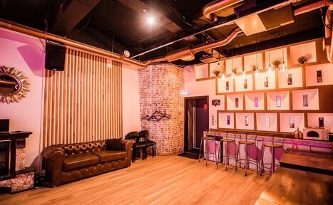 Арендовать LoftForParty. Бэкстейдж лофт . Лофт-пространство с барной зоной и кирпичной стеной. Диван и столы из темного дерева, барные стулья на высоких ножках. Пол из светлого дерева. Рейки на стене, декоративные лампы на потолке, открытые вентиляционные трубы. Теплое освещение.