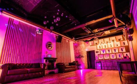 Арендовать LoftForParty. Бэкстейдж лофт . Клубное помещение с фиолетовым освещением, черным потолком и открытыми трубами. Два кожаных дивана у стен, барная стойка с высокими табуретами и полки. Деревянный пол, зеркальные шары на потолке, кирпичная стена, декоративные панели.