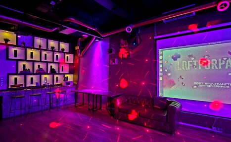 Арендовать LoftForParty. Бэкстейдж лофт . Тёмный лофт с неоновым освещением, барной стойкой и высокими стульями. На стене большой экран. Кожаный диван стоит у стены. Декоративные растения на полках. Потолочные трубы, деревянный пол. Надпись на экране: "Loft-пространство для вечеринок".