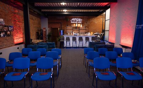 Арендовать MPSellers(МПСеллерс). Просторный зал для мероприятий. Пространство с кирпичными стенами, ряды синих стульев, барная стойка с неоновой надписью, сцена с микрофонами, мягкое боковое освещение, транспортная металлическая конструкция под потолком.