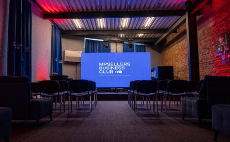 Арендовать MPSellers(МПСеллерс). Просторный зал для мероприятий. Аудитория с экраном, креслами в ряд, стульями. Кирпичные стены, потолок с металлическими балками. Синий и красный свет. Занавески на окнах. Монитор с текстом "MPSELLERS BUSINESS CLUB". Декоративные элементы на стенах.