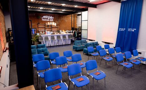 Арендовать MPSellers(МПСеллерс). Просторный зал для мероприятий. Просторное помещение с кирпичными стенами и металлическими балками. Синие стулья в ряд перед диванами. Барная стойка с высокими стульями. Неоновые и подвесные светильники. Ковер на полу. Цвета: синий, белый, кирпичный. Надпись на стене: "MPS club".
