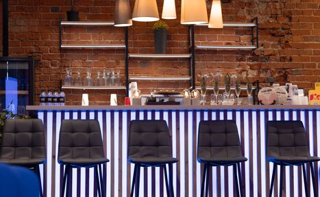 Арендовать MPSellers(МПСеллерс). Просторный зал для мероприятий. Барная стойка с высокими стульями, кирпичная стена, полки с бокалами и бутылками, освещение лампами подвесного типа, светодиодная подсветка синего цвета, металлические и стеклянные элементы, декоративное растение.