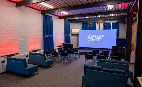 Арендовать MPSellers(МПСеллерс). Просторный зал для мероприятий. Пространство с рядами стульев перед большим экраном. Слева расположены синие диваны. Белые кирпичные стены, на полу серый ковролин. Яркие синие шторы на окнах. Подвесной потолок с металлическими панелями. Надпись на экране: "MPSELLERS BUSINESS CLUB".