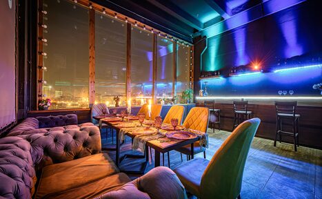 Арендовать Лофт ZUM(Зум). Пространство с мягкими диванами и стульями вокруг обеденного стола, сервированного посудой. Большие окна со шторками, светодиодная подсветка на стенах. Потолок тёмный, пол плиточный. Декоративные огни вдоль стены, барная стойка со стульями.