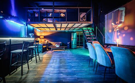 Арендовать Лофт ZUM(Зум). Лофт-пространство с барной стойкой и стульями слева, зоной для отдыха с диваном и столиком справа, и лестницей на второй уровень. Синий и фиолетовый свет, деревянный пол, изображение микрофона на стене, стулья серых оттенков.