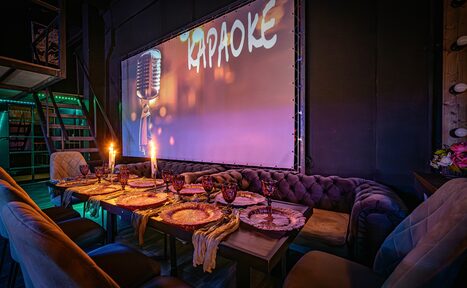 Арендовать Лофт ZUM(Зум). Караоке-зал с обеденным столом, сервированным на шесть персон. Стена с большим экраном, на котором изображен микрофон и слово "Караоке". Диван и стулья вдоль стола, свечи, мягкое освещение. Тёмные стены и пол, деревянные панели.