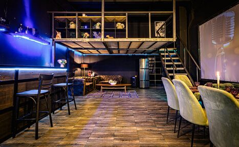 Арендовать Лофт ZUM(Зум). Лофт-пространство с мезонином и деревянной лестницей, креслами и высокими стульями, деревянный пол, неоновая подсветка, темные стены, зона отдыха с диваном, журнальный столик, холодильник, настольная лампа, декоративные элементы на втором уровне.