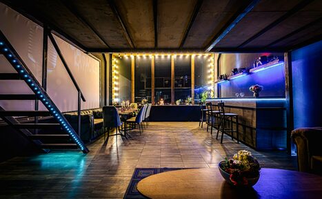 Арендовать Лофт ZUM(Зум). Лофт с деревянным полом и открытыми потолочными балками. Лестница с подсветкой. Большие окна с гирляндой ламп. Барная стойка с подсветкой, высоким барным стульями. Диван вдоль стены и стол с цветочным декором. Тёмные и синие цвета.