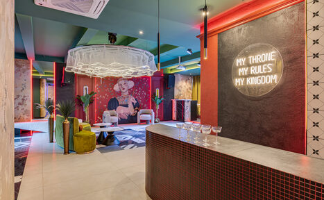 Арендовать Стильное пространство Астрид. Современное пространство с барной стойкой, неоновым знаком "MY THRONE MY RULES MY KINGDOM", мягкой зоной с креслами и журнальным столиком, зелеными растениями, яркими стенами и декоративными элементами. Потолок светлый, плитка на полу.